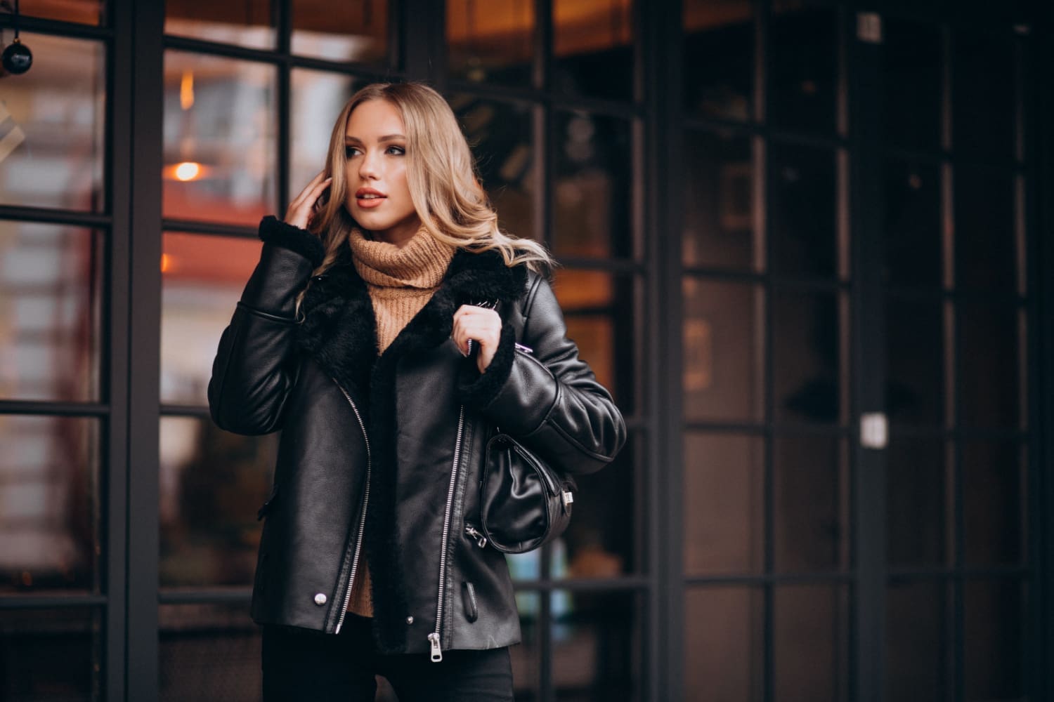 A woman in a leather jacket and brown turtleneck sweater stands outside, talking on her phone. She is in front of a glass window with a black grid frame.