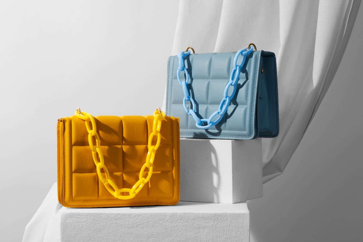 Two quilted handbags on display: one yellow with a chunky yellow chain, and one teal with a blue chain, both placed on white pedestals.