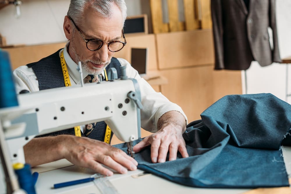 An older man with glasses uses a sewing machine to stitch blue fabric in a tailor's workshop. Measuring tape hangs around his neck.